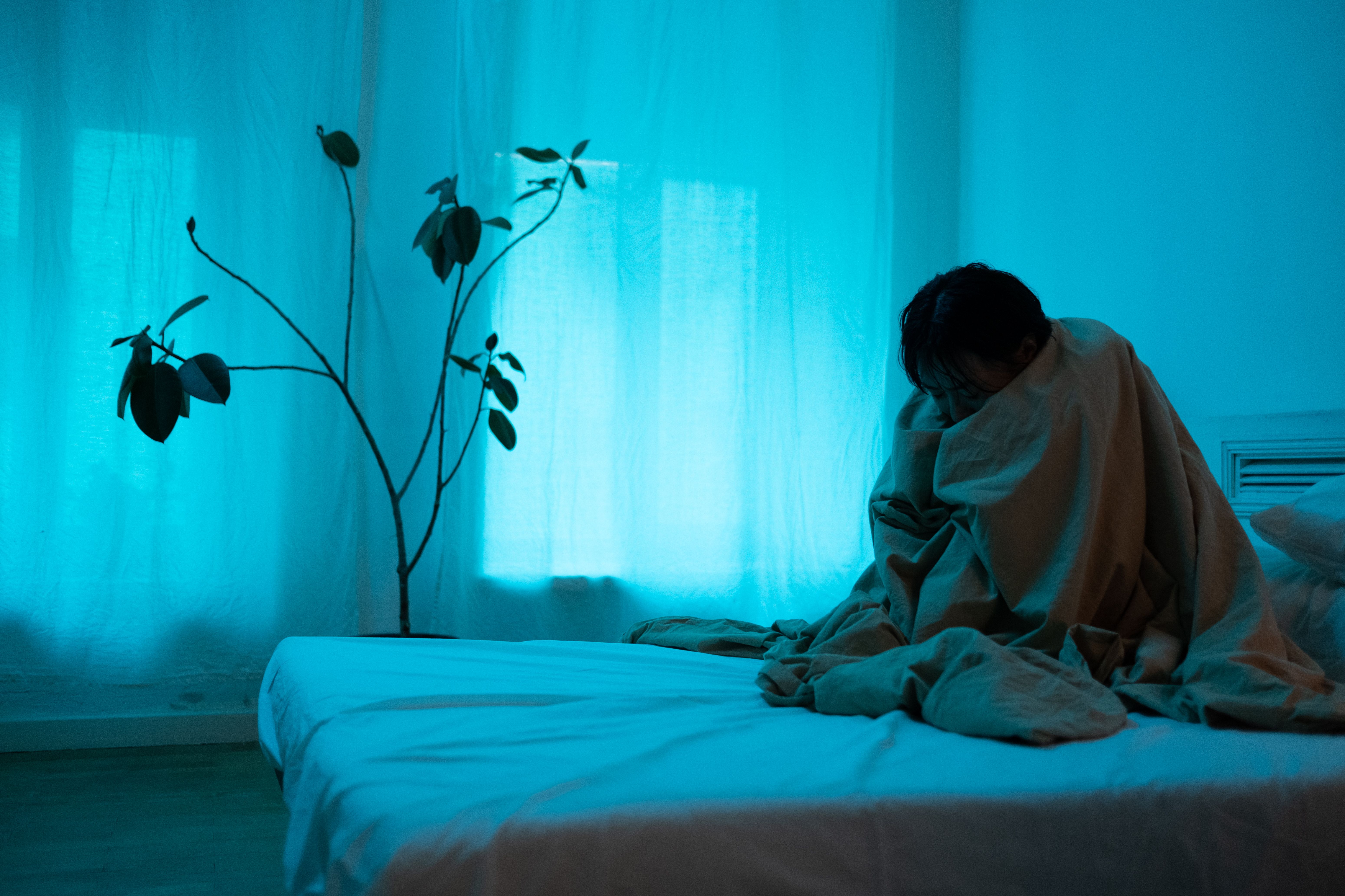 A young person sits alone on a bed wrapped in a blanket, conveying isolation and emotional distress.